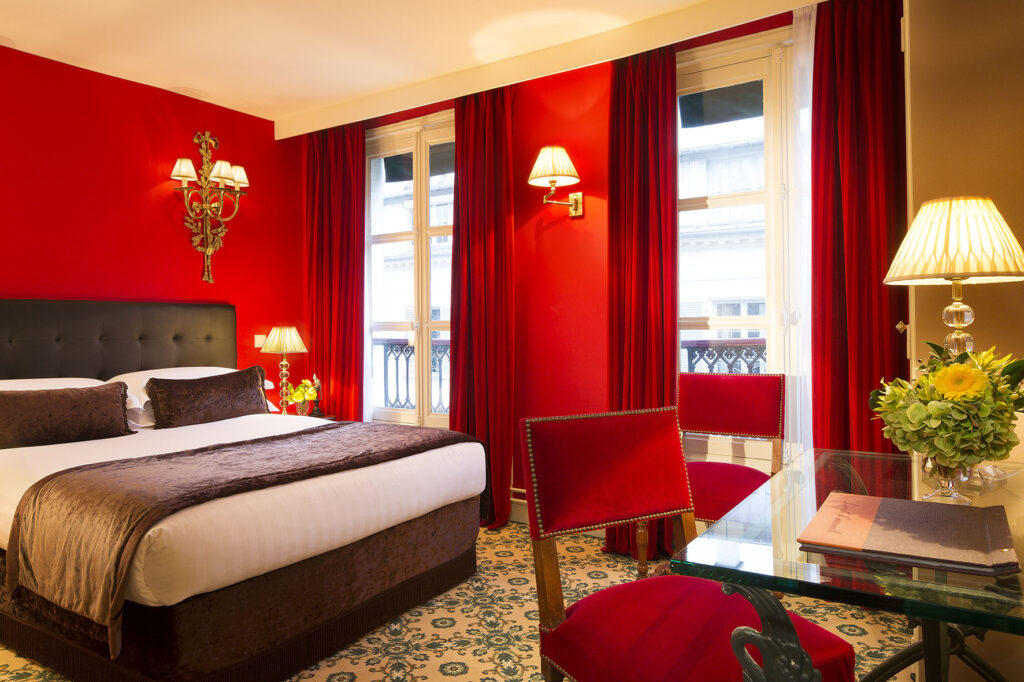 Photo d'une chambre - murs rouges, lit double avec coussins et plaid marrons, chaises rouges, bureau et deux grandes fenêtres sur la ville - hôtel dans le meilleur quartier de Paris : Hôtel des 2 Continents Paris 6