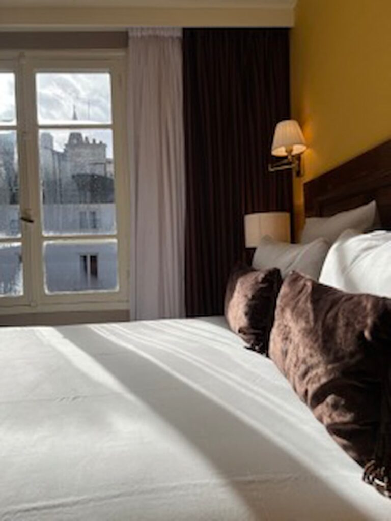 Hôtel Roland Garros Paris: Voici une chambre cosy à l'Hôtel des 2 Continents. Elle dispose d'un lit double avec des draps blanc. Les murs sont jaunes et les rideaux marrons. Cette chambre a la chance d'offrir une vue sur le cloché de l'église Saint-Germain-des-Prés. 