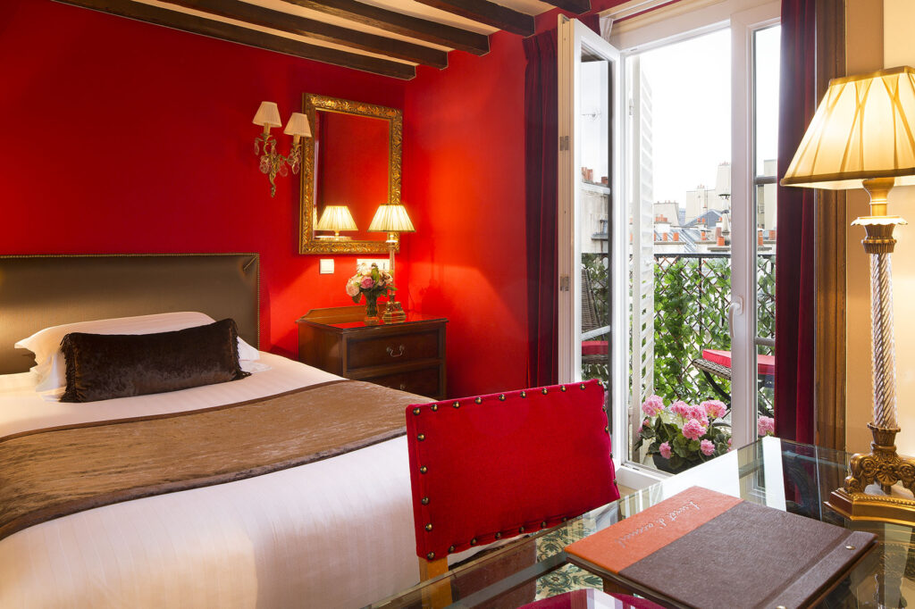 Hotel for Roland Garros Paris: We discover our superior room with a balcony. The predominant color is red (walls, chairs). Lamps are beige and wooden beams shape the ceiling. On our right we see the balcony and a magnificent view of the Parisian rooftops.