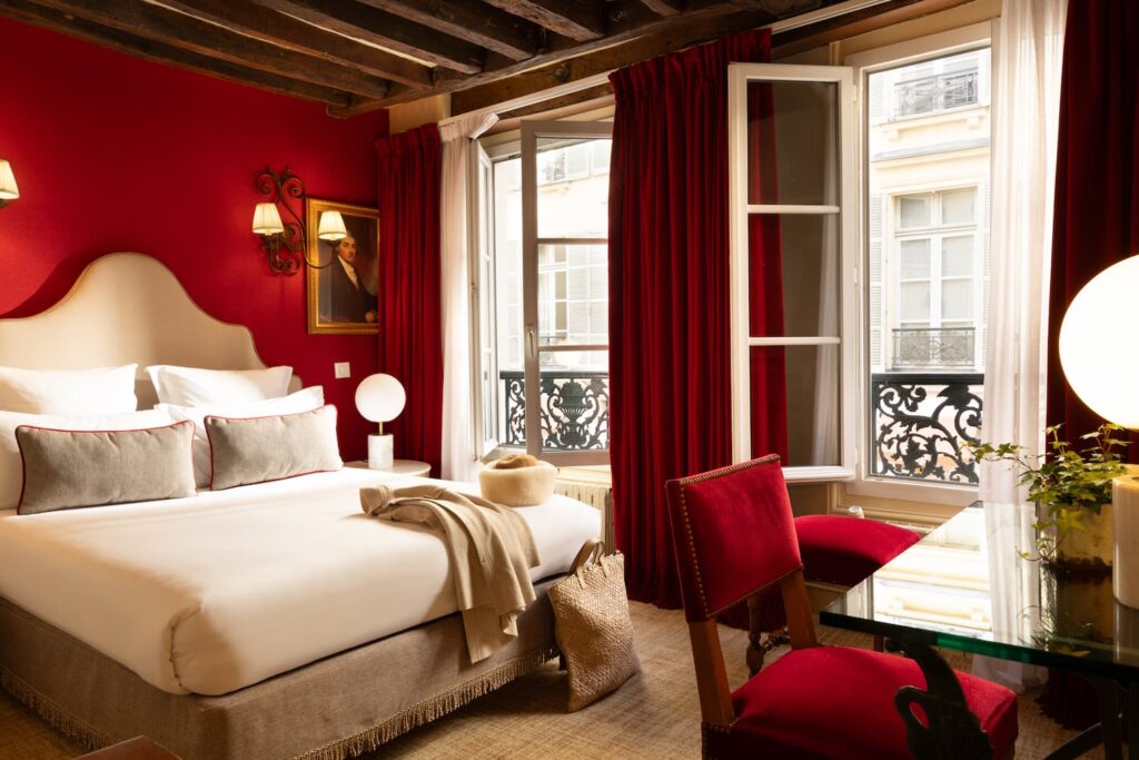 chambre avec fenêtres ouvertes sur la rue, lit blanc, manteau et chapeau beiges posés sur le lit et tissu rouge | soirée romantique à paris et à l'Hôtel des 2 Continents
