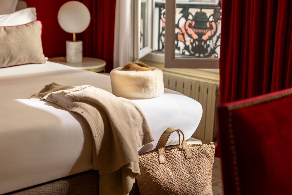 manteau et chapeau beiges posés sur un lit blanc, sac beige au pied du lit, fenêtre ouverte, rideau et tissu rouges | soirée romantique à paris et à l'Hôtel des 2 Continents