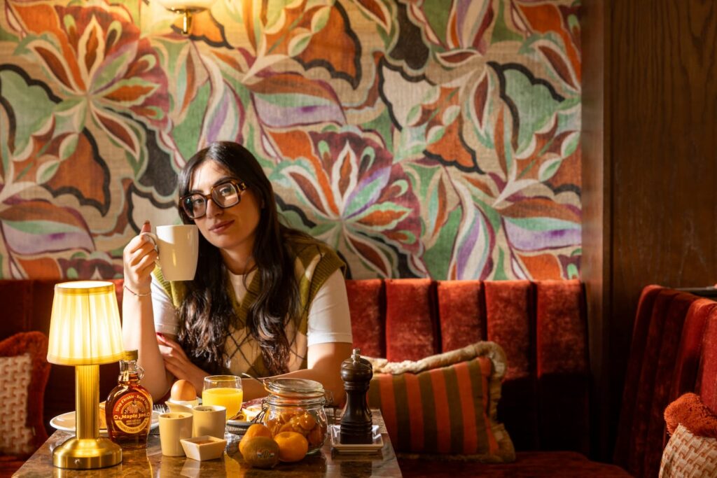 jeune femme assise à sa table de petit-déjeuner, avec une tasse dans la main, une lampe sur la table, des fruits et du sirop d'érable devant elle | petit-déjeuner healthy paris hotel des 2 Continents